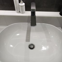 Czysty Dom - Biała, owalna umywalka z chromowaną armaturą i zestawem kosmetyków Vivian Gray z kryształkami na blacie, na tle ciemnej ściany.