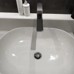 Biała, owalna umywalka z chromowaną armaturą i zestawem kosmetyk&oacute;w Vivian Gray z kryształkami na blacie, na tle ciemnej ściany.