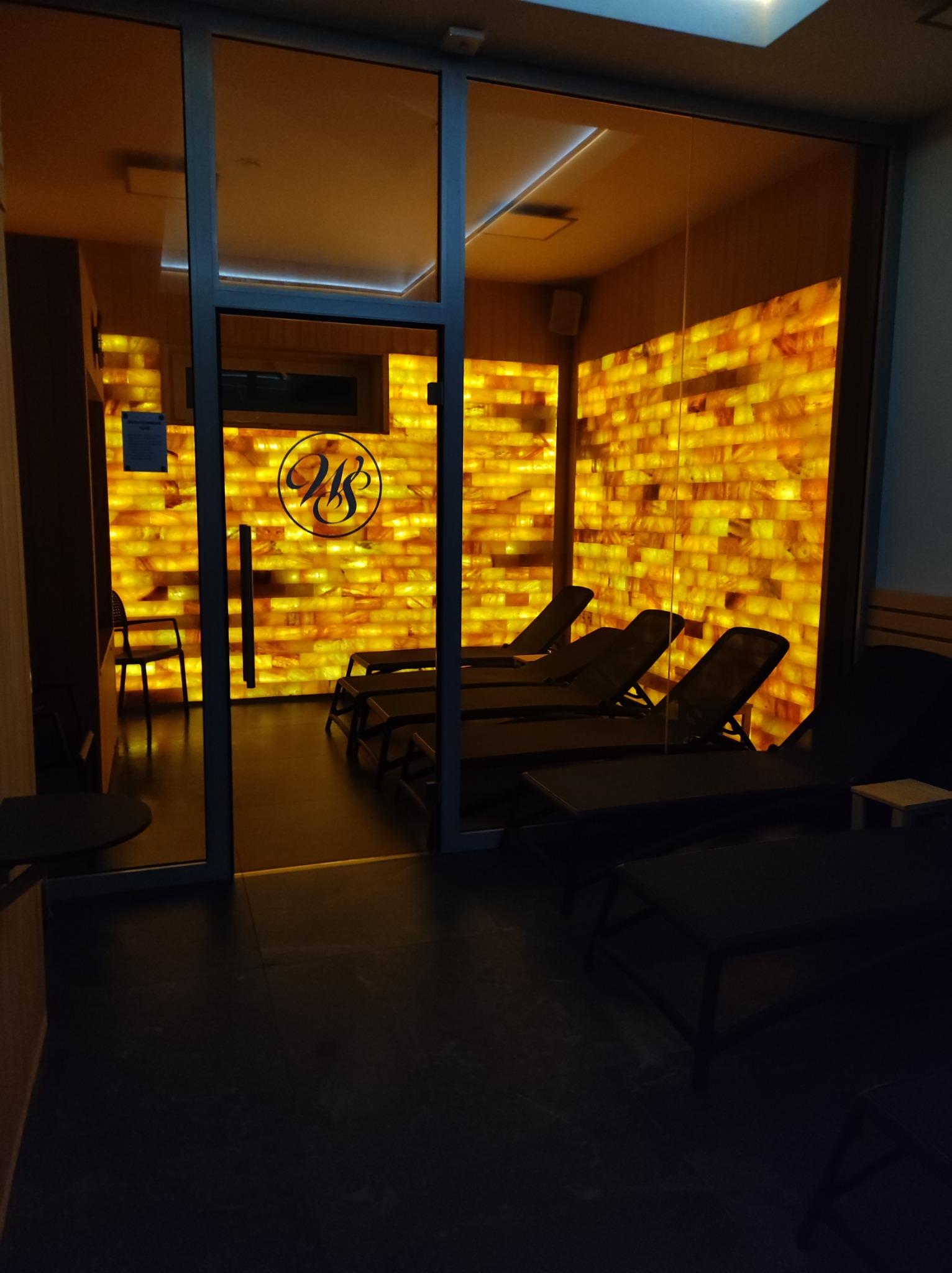 Wnętrze relaksacyjne z ścianą z cegieł solnych, leżakami i logo na szklanej ścianie, oświetlone ciepłym światłem.