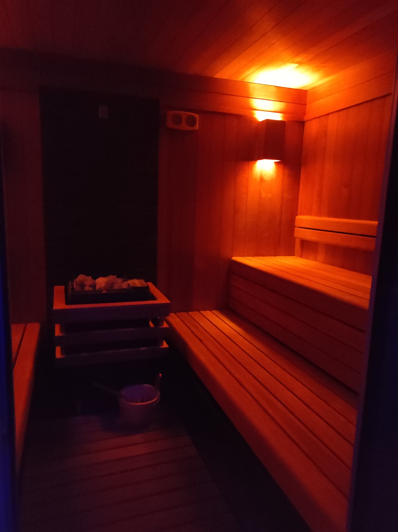 Wnętrze sauny z drewnianymi ławkami, piecem z kamieniami i oświetleniem w ciepłej tonacji, widziane z perspektywy wejścia.