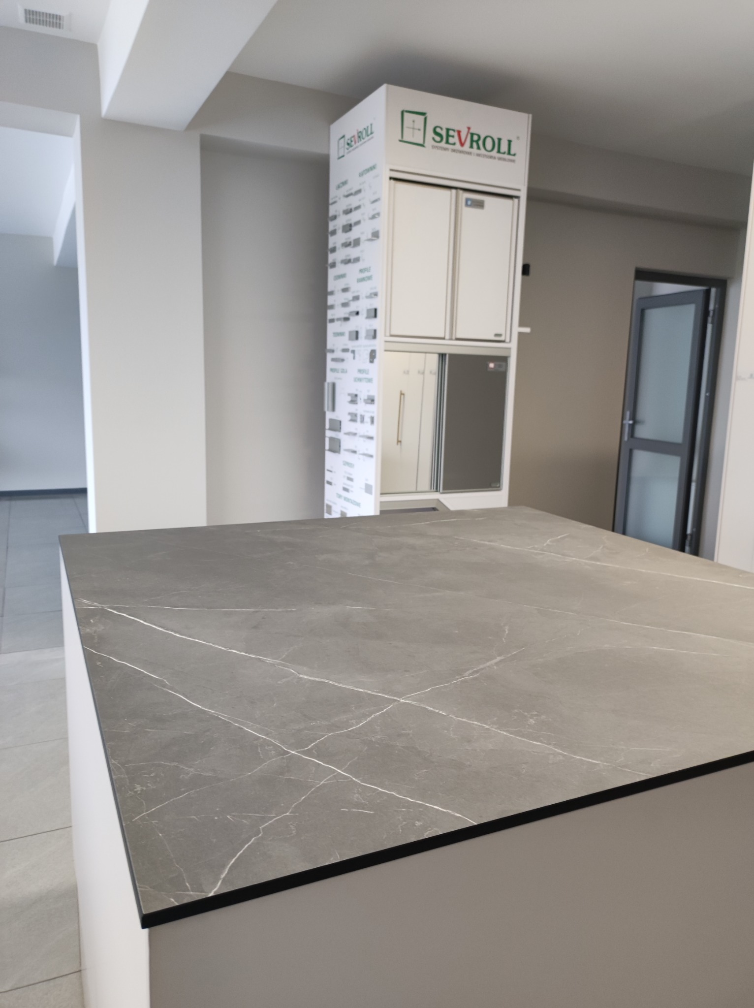 Szara, marmurkowa powierzchnia blatu kuchennego na tle ekspozycji szafek firmy Sevroll i otwartego pomieszczenia.