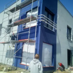 Prace na zewnątrz budynku: robotnicy na rusztowaniu i przy fundamentach, elewacja w trakcie malowania na granatowo i szaro, okna zabezpieczone folią.