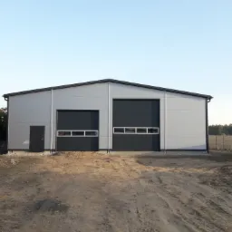 Nowo wybudowana hala z płyt warstwowych, z dwiema bramami segmentowymi i drzwiami wejściowymi, widoczna sterta kostki brukowej przed budynkiem.