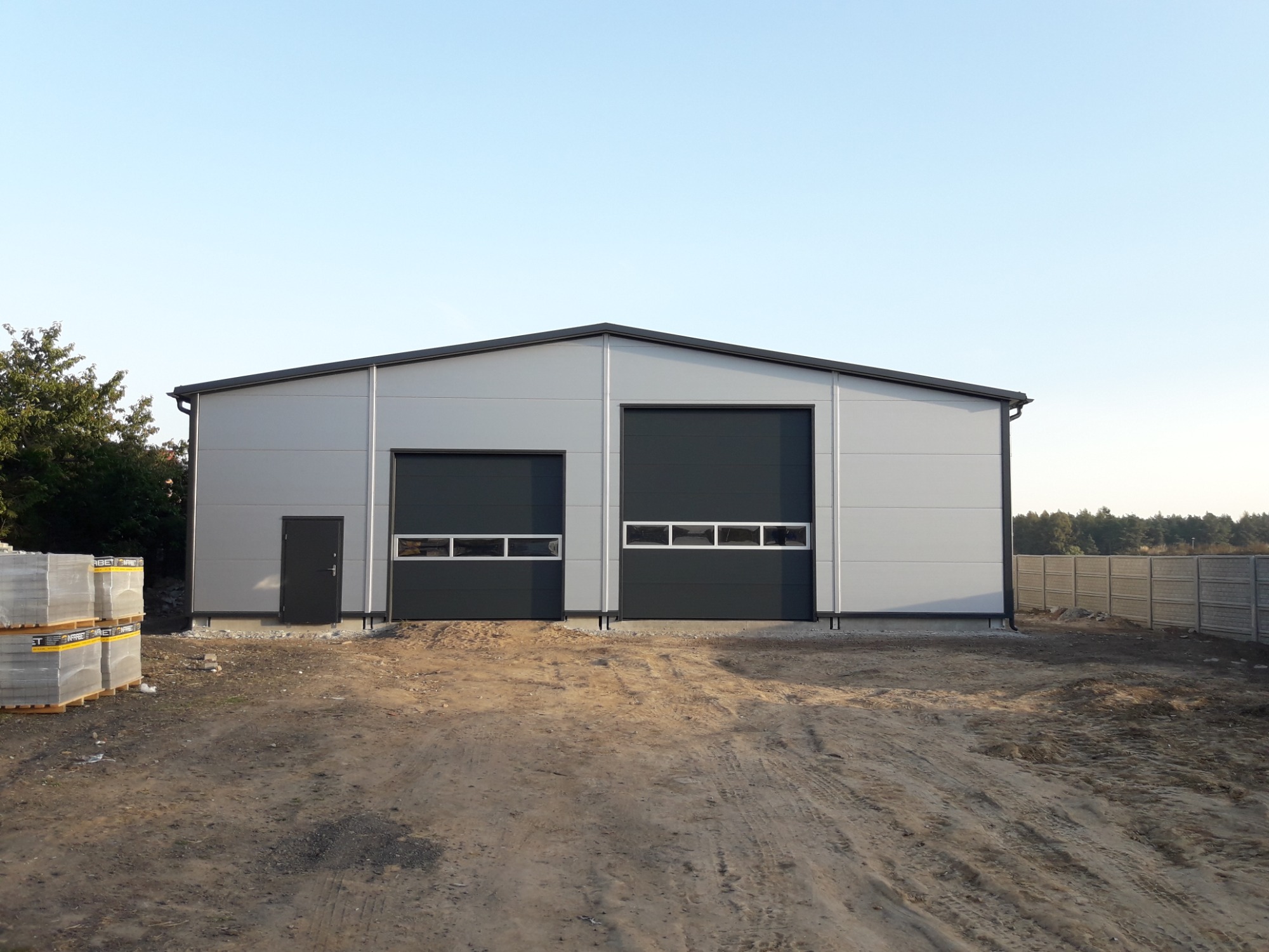 Nowo wybudowana hala z płyt warstwowych, z dwiema bramami segmentowymi i drzwiami wejściowymi, widoczna sterta kostki brukowej przed budynkiem.