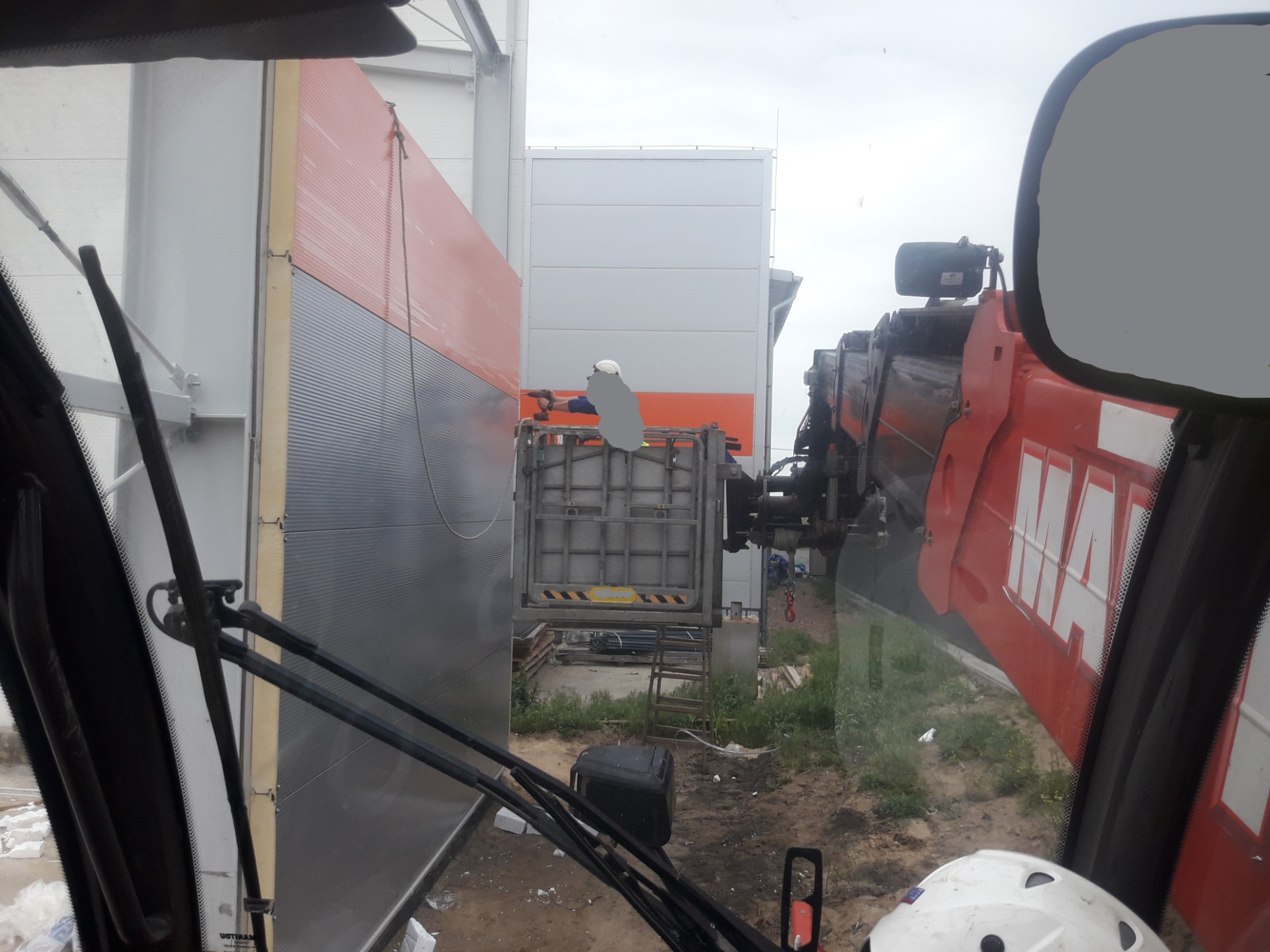 Pracownik w kasku, z podnośnika, montuje panel warstwowy na ścianie hali, widok z kabiny dźwigu, w tle elementy konstrukcji stalowej.