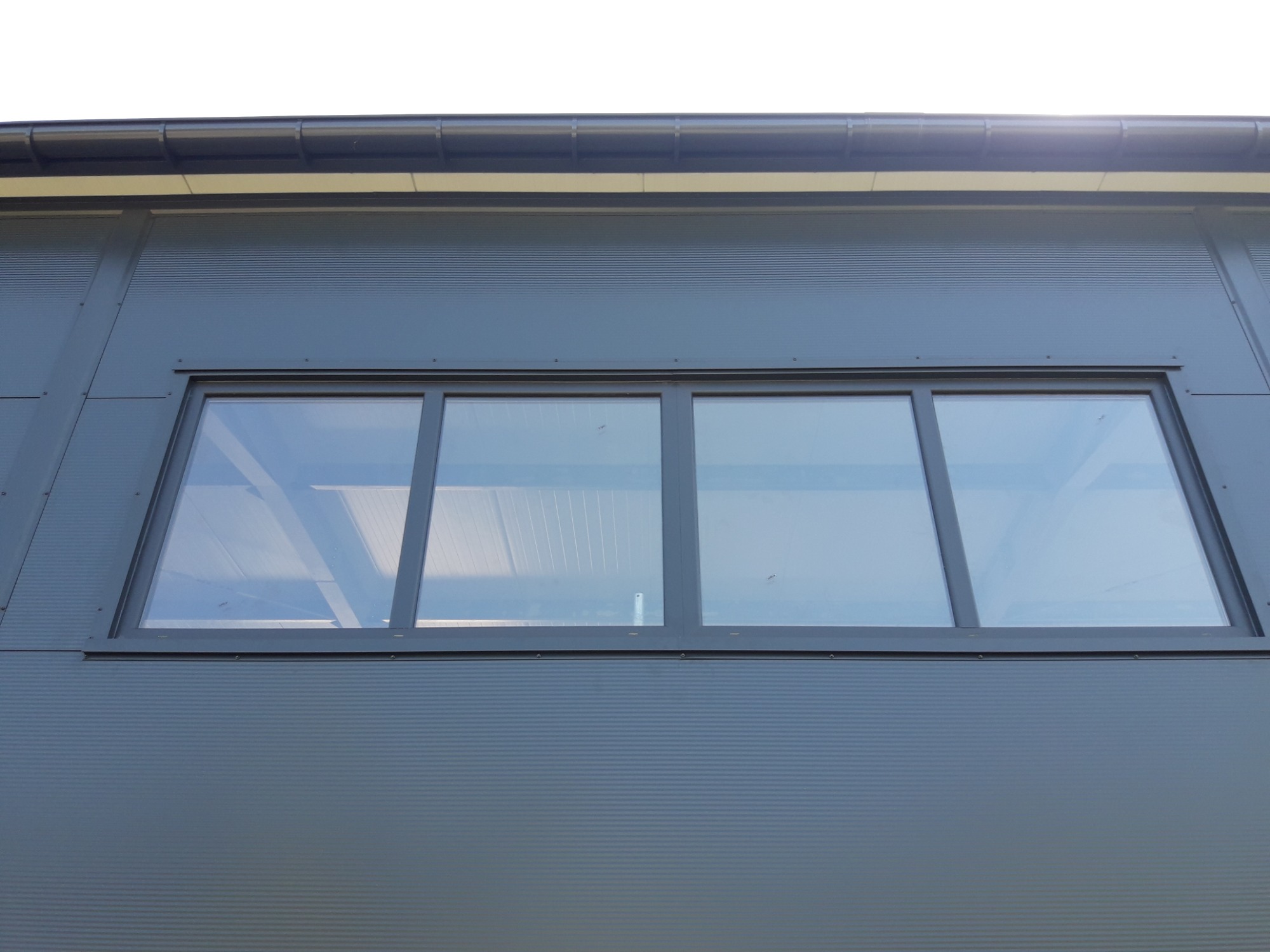 Szara ściana hali w Lubaszu, wykonana z płyty warstwowej z zamontowanym oknem trzyskrzydłowym z ciemną ramą.