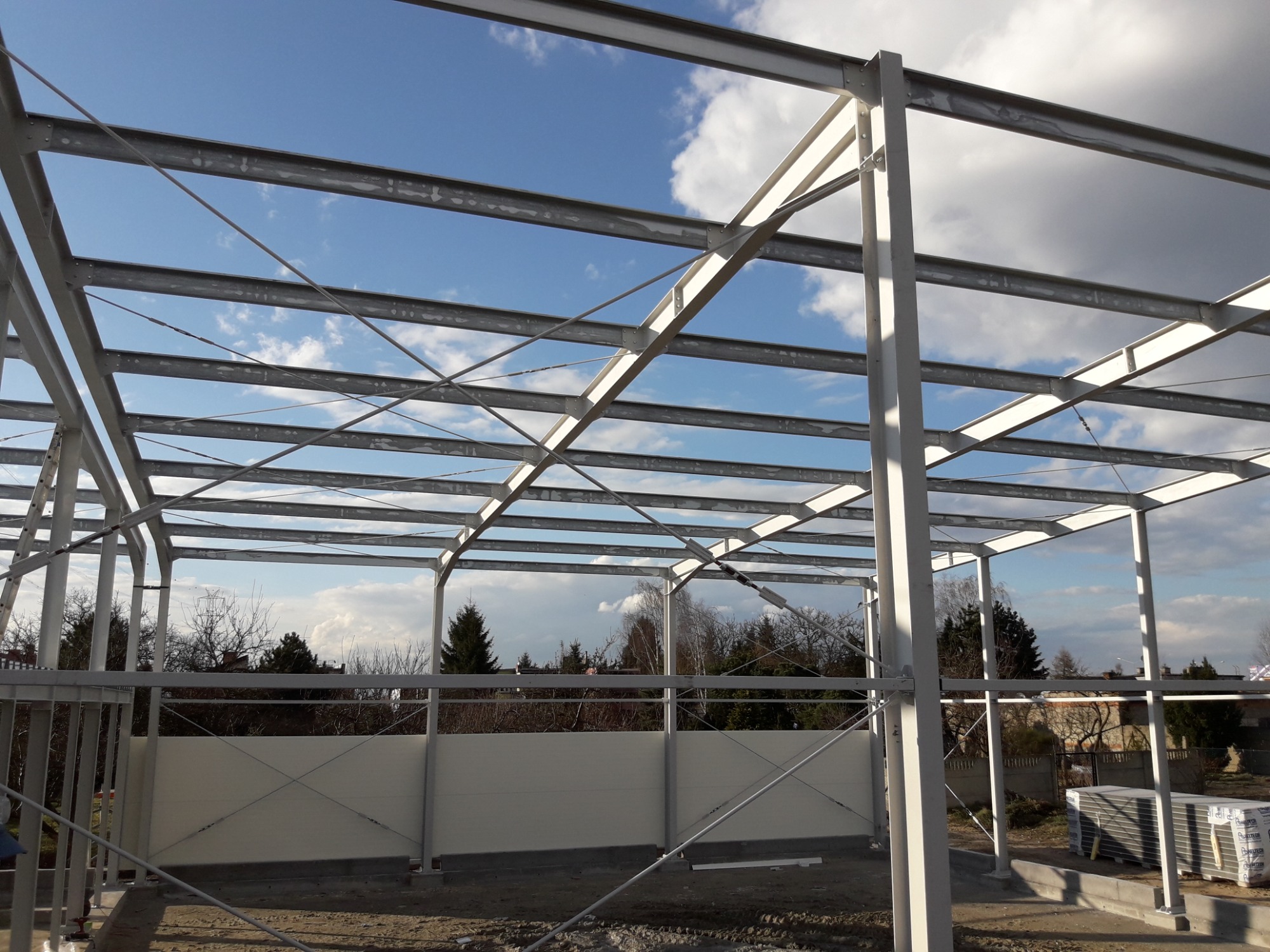 Konstrukcja stalowa hali w trakcie budowy, widoczne białe słupy i poprzeczki, w tle błękitne niebo z chmurami oraz częściowo zamontowane białe płyty warstwowe.
