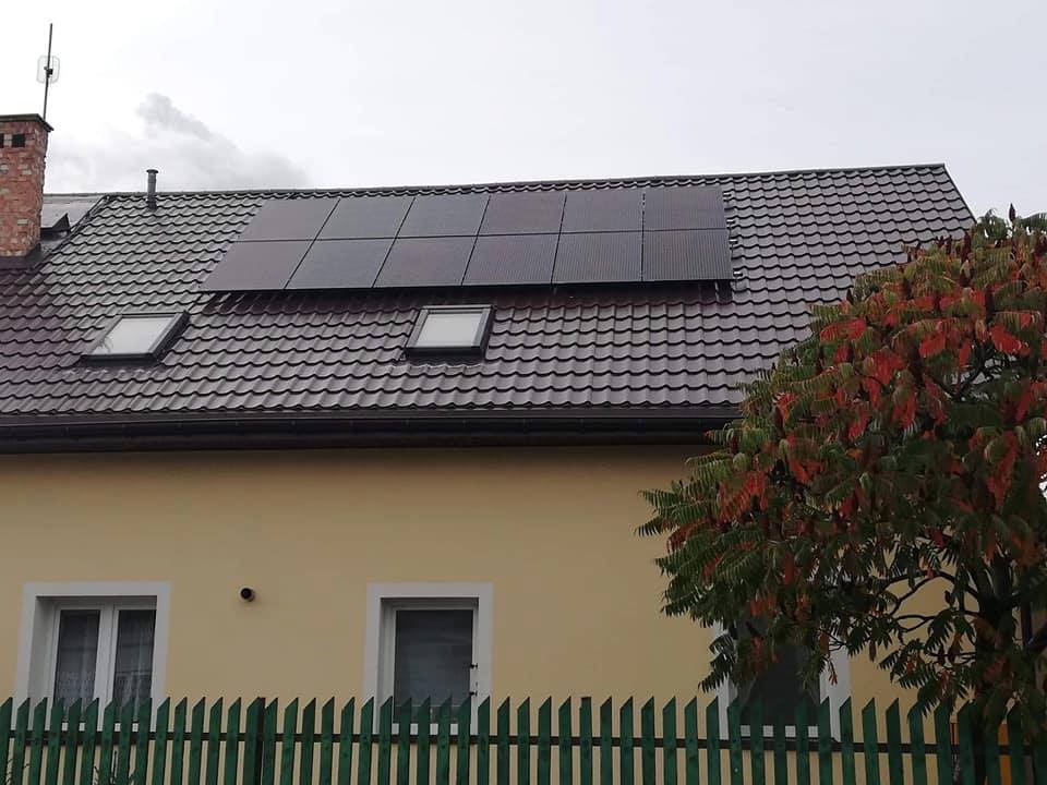 Panele słoneczne na brązowym dachu domu z oknami dachowymi, widok zza zielonego płotu i drzewa z czerwieniejącymi liśćmi.