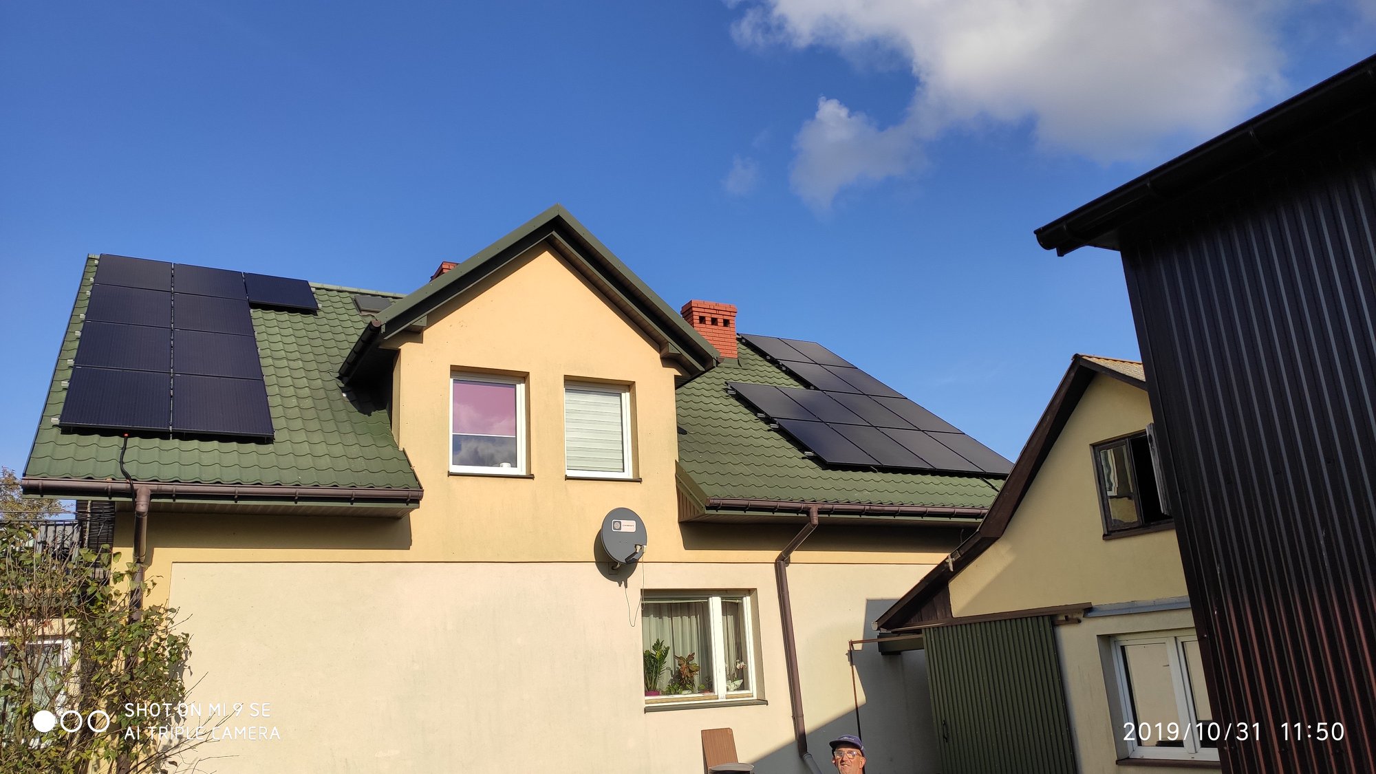 Panele słoneczne na zielonym dachu dwupiętrowego domu z beżową elewacją, widoczne satelita, okna z roletami i mały fragment osoby w dolnym rogu.