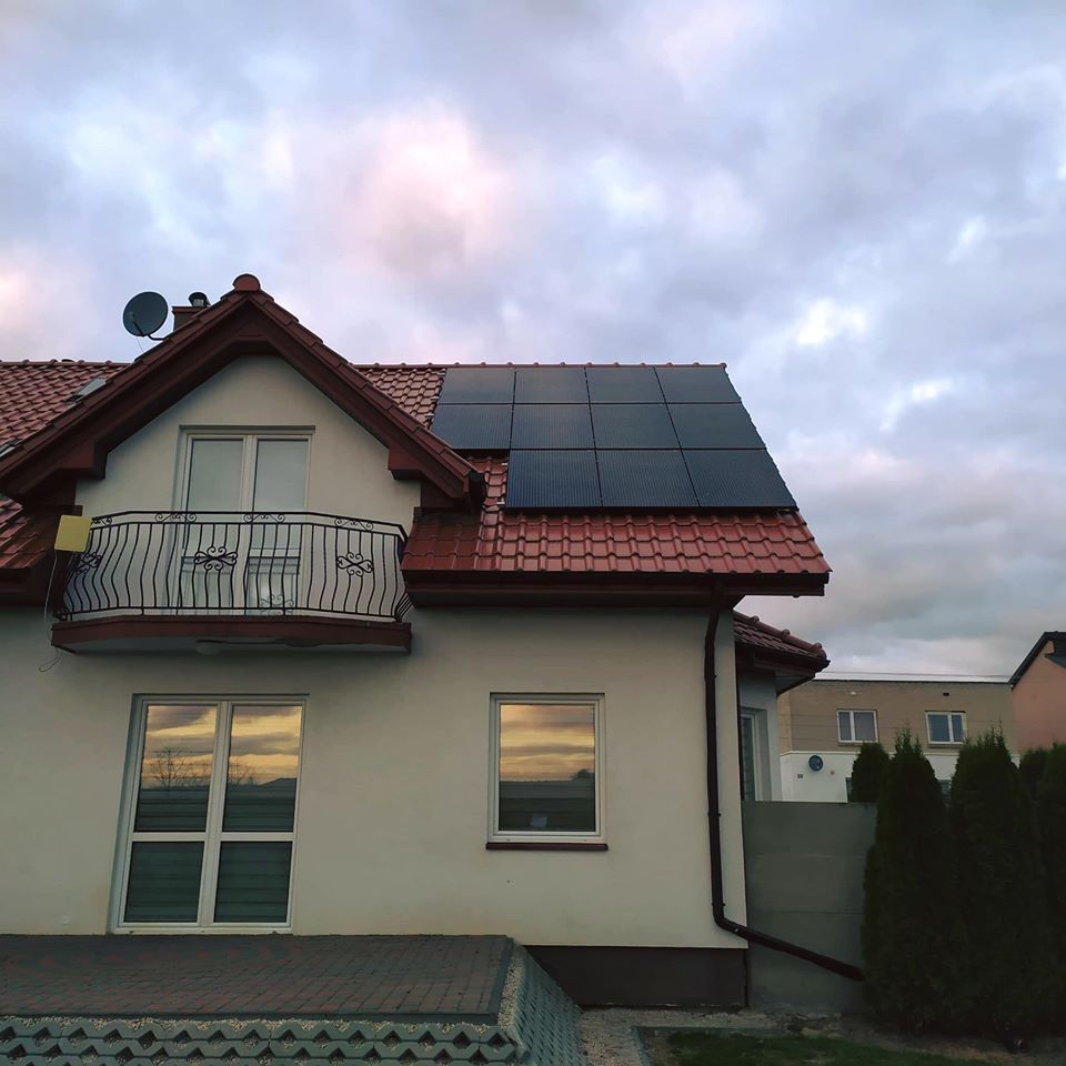 Dom jednorodzinny z panelami słonecznymi na czerwonym dachu, widoczne odbicie zachodu słońca w oknach i balkon z ozdobną balustradą.