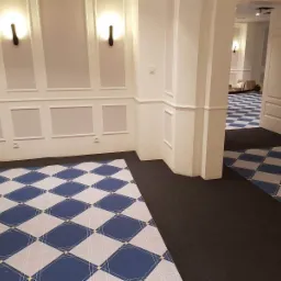 Wnętrze z nową wykładziną w romby w odcieniach bieli i granatu, kontrastującą z ciemną, jednolitą wykładziną w przejściu, białe ściany z dekoracyjnymi panelami i czarne kinkiety