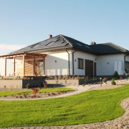 Parterowy dom z panelami solarnymi na dachu, jasną elewacją i drewnianą pergolą. Uwagę zwraca zadbany trawnik i ścieżka z kostki brukowej.