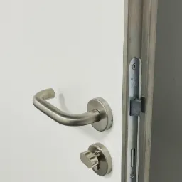 Białe skrzydło z klamką i zamkiem w kolorze satynowego niklu, widoczny mechanizm zamka na krawędzi ościeżnicy.