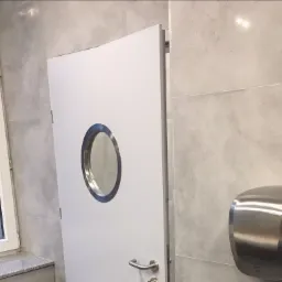 Białe drzwi wewnętrzne z okrągłym okienkiem ze stali nierdzewnej, zamontowane w łazience wykończonej szarymi płytkami, obok suszarka do rąk.