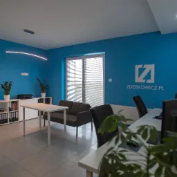 Wnętrze biura firmy Zdzislowicz.pl z niebieskimi ścianami, logo firmy na ścianie, biurkami, krzesłami, minimalistycznym oświetleniem i roślinami doniczkowymi.