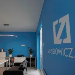 Błękitne biuro z logo Zdzislowicz.pl na ścianie, minimalistyczne wnętrze z sofą, stołem i roślinami doniczkowymi, podłużna lampa sufitowa.