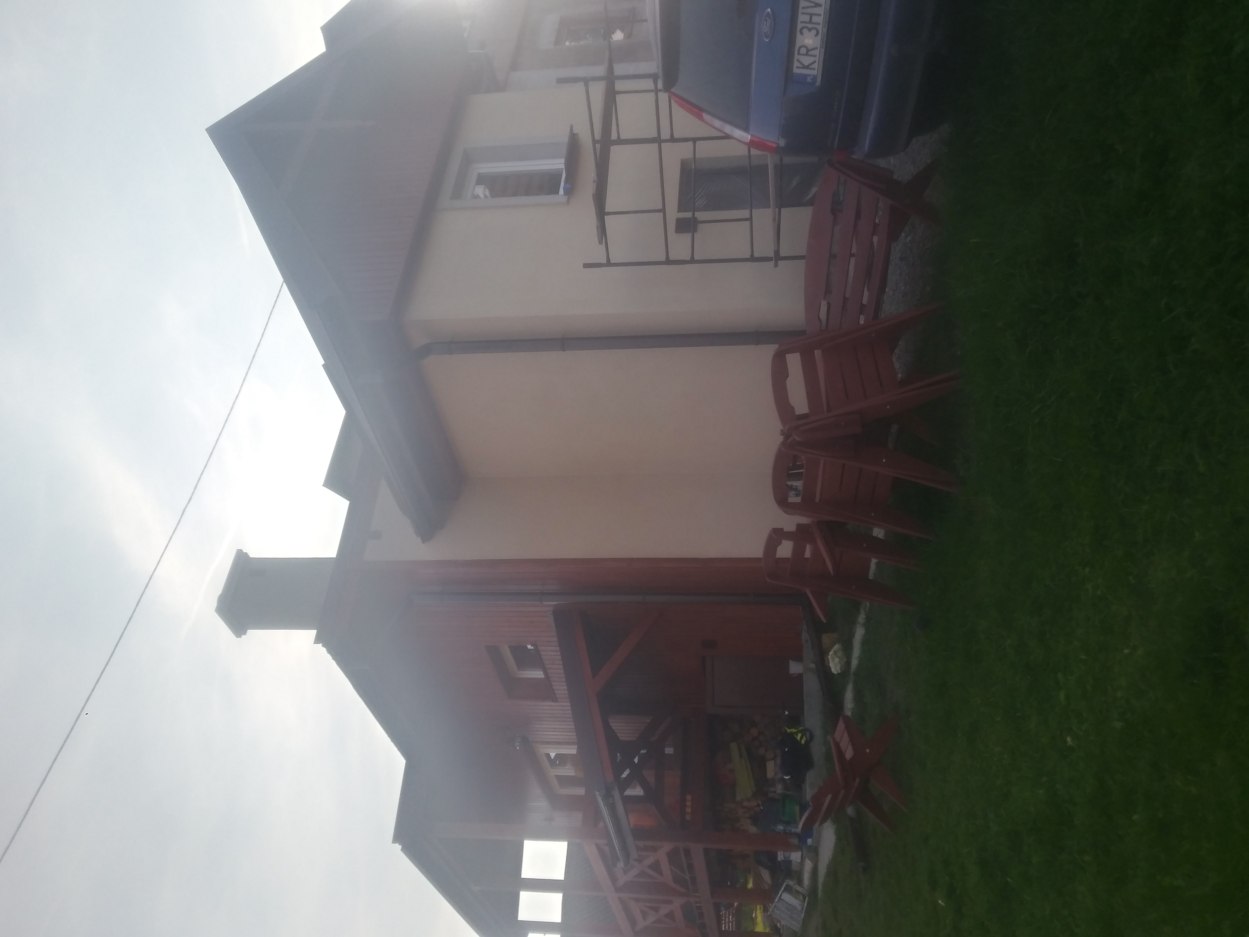 Ocieplony dom jednorodzinny z jasną elewacją, widoczne rusztowanie przy ścianie, ułożone krzesła ogrodowe i zaparkowany samochód.