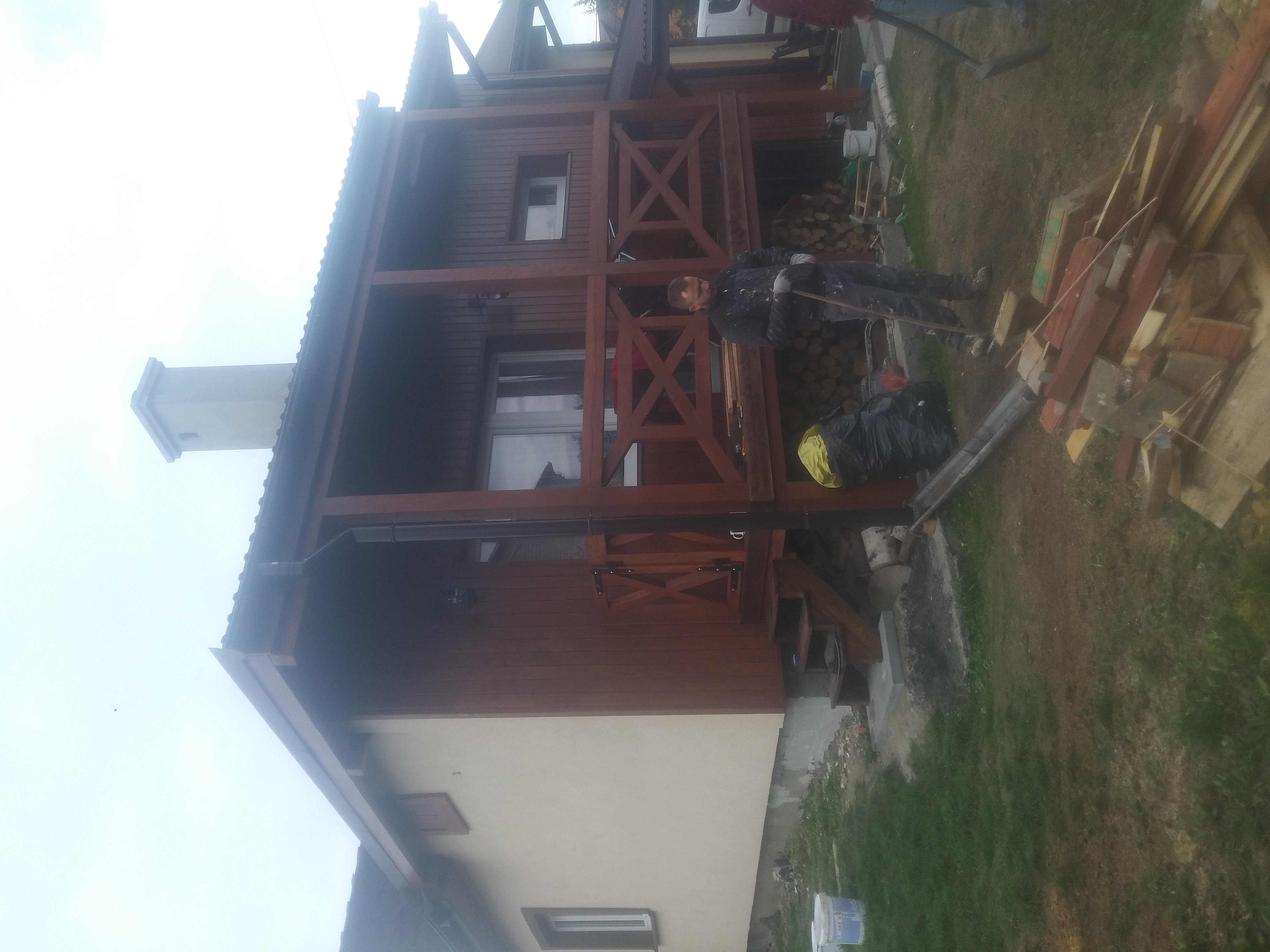 Dwukondygnacyjny dom z drewnianą konstrukcją werandy, pracownik w kombinezonie roboczym z narzędziem przy stercie drewna i workach na śmieci, widok na elewację i komin.