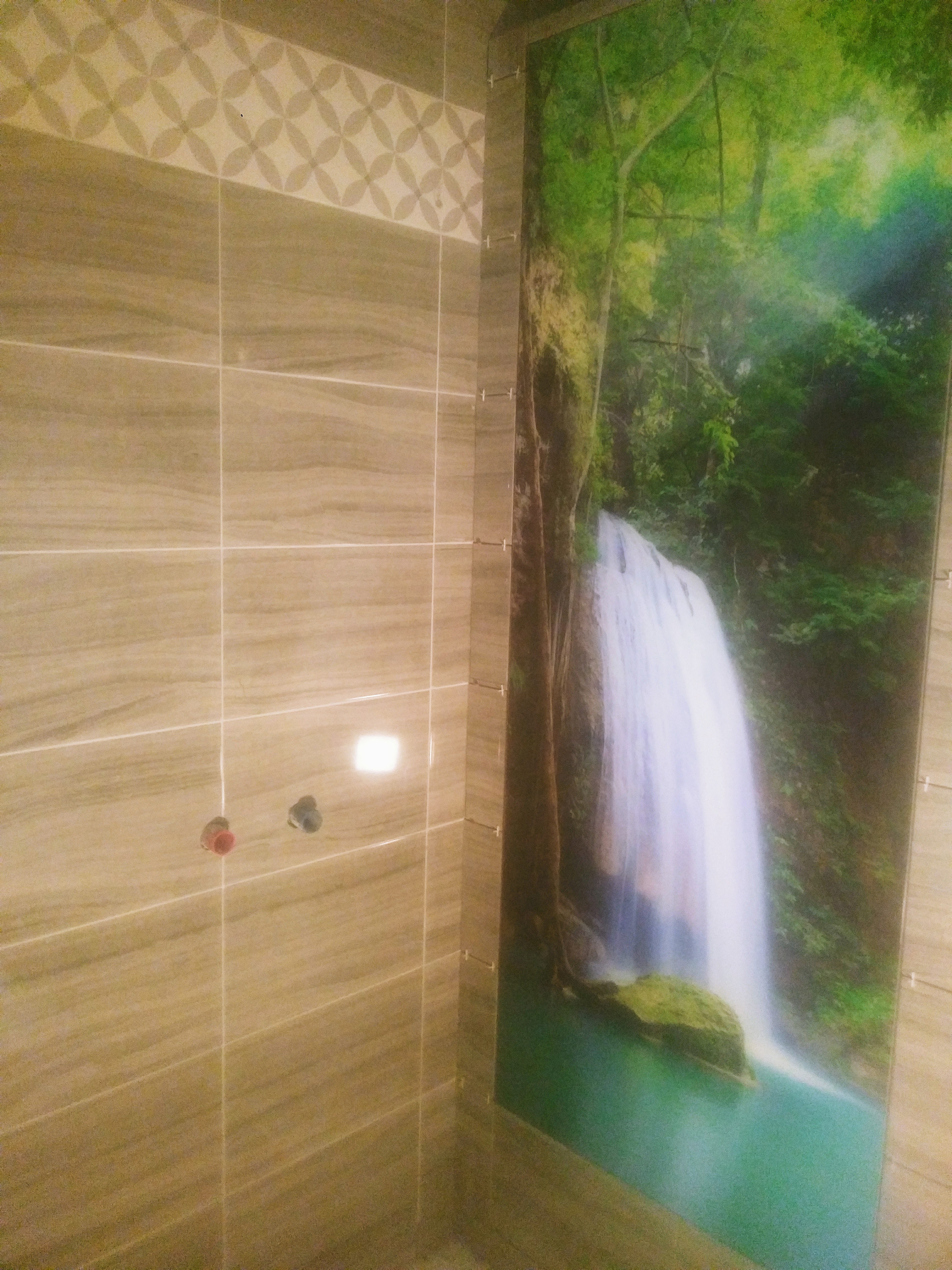 Ściana łazienki wyłożona beżowymi płytkami z dekoracyjnym pasem i fototapetą z motywem wodospadu w lesie tropikalnym, widoczne zawory instalacji wodnej.