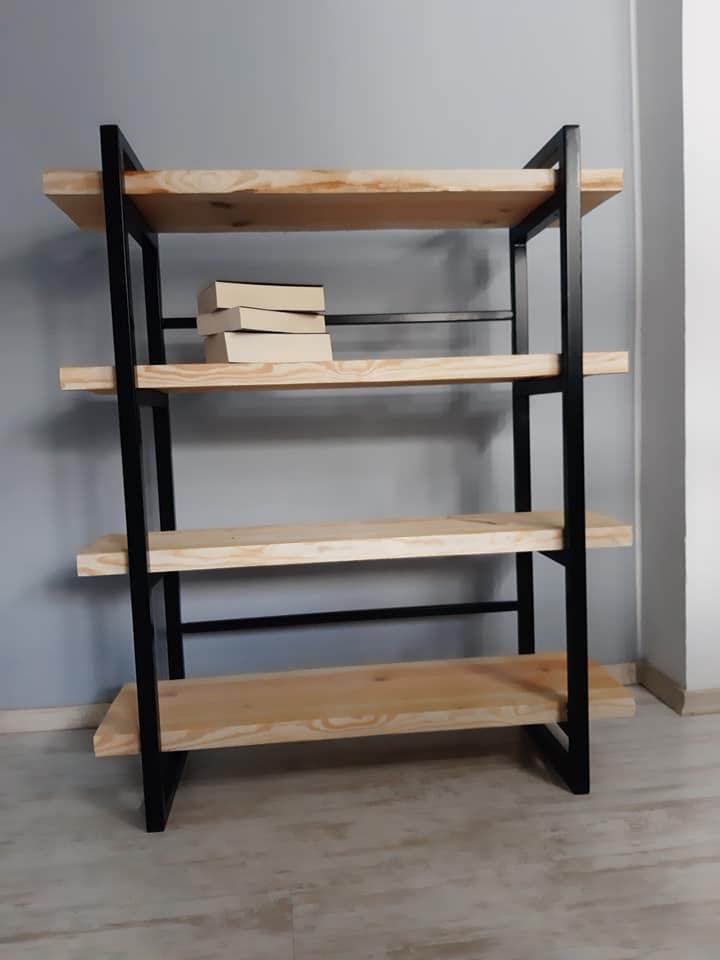 Regał z drewnianymi półkami i czarną metalową ramą, trzy książki ułożone na drugiej półce od góry, szary mur w tle.