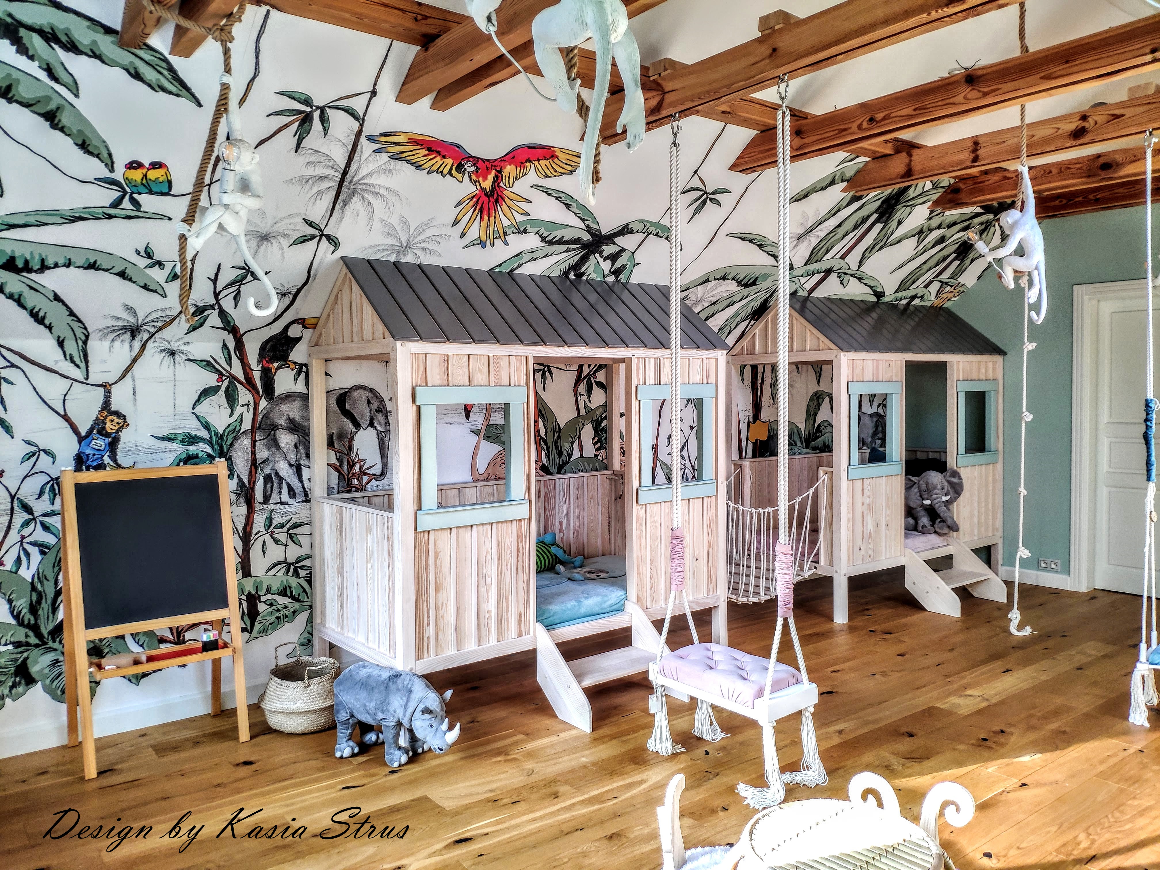 Pokój dziecięcy z dwoma drewnianymi domkami-łóżkami połączonymi mostkiem, huśtawka na linach, ściana z motywem tropikalnym, drewniana podłoga i dekoracje w postaci małp i nosorożca.