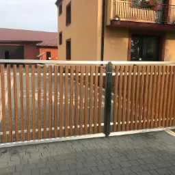 Nowoczesna brama wjazdowa z drewnianymi lamelami i metalową ramą, widok na dom z balkonem i garaż w tle.