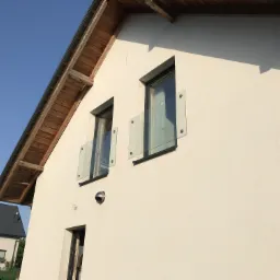 Nowoczesna elewacja domu z jasnego tynku z drewnianym okapem i szklanymi balustradami przy oknach na piętrze.