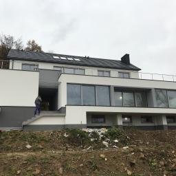 FrezDruk - Nowoczesny dom z dużymi oknami, metalową balustradą na dachu i osobą wchodzącą po schodach zewnętrznych, teren wokół domu w trakcie prac wykończeniowych.