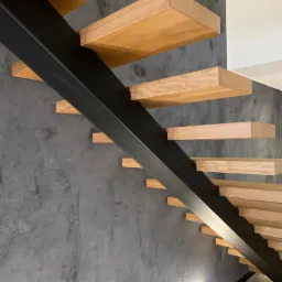 Nowoczesne schody z drewnianymi stopniami i czarną, metalową konstrukcją nośną, zamontowane przy szarej ścianie o teksturze betonu.