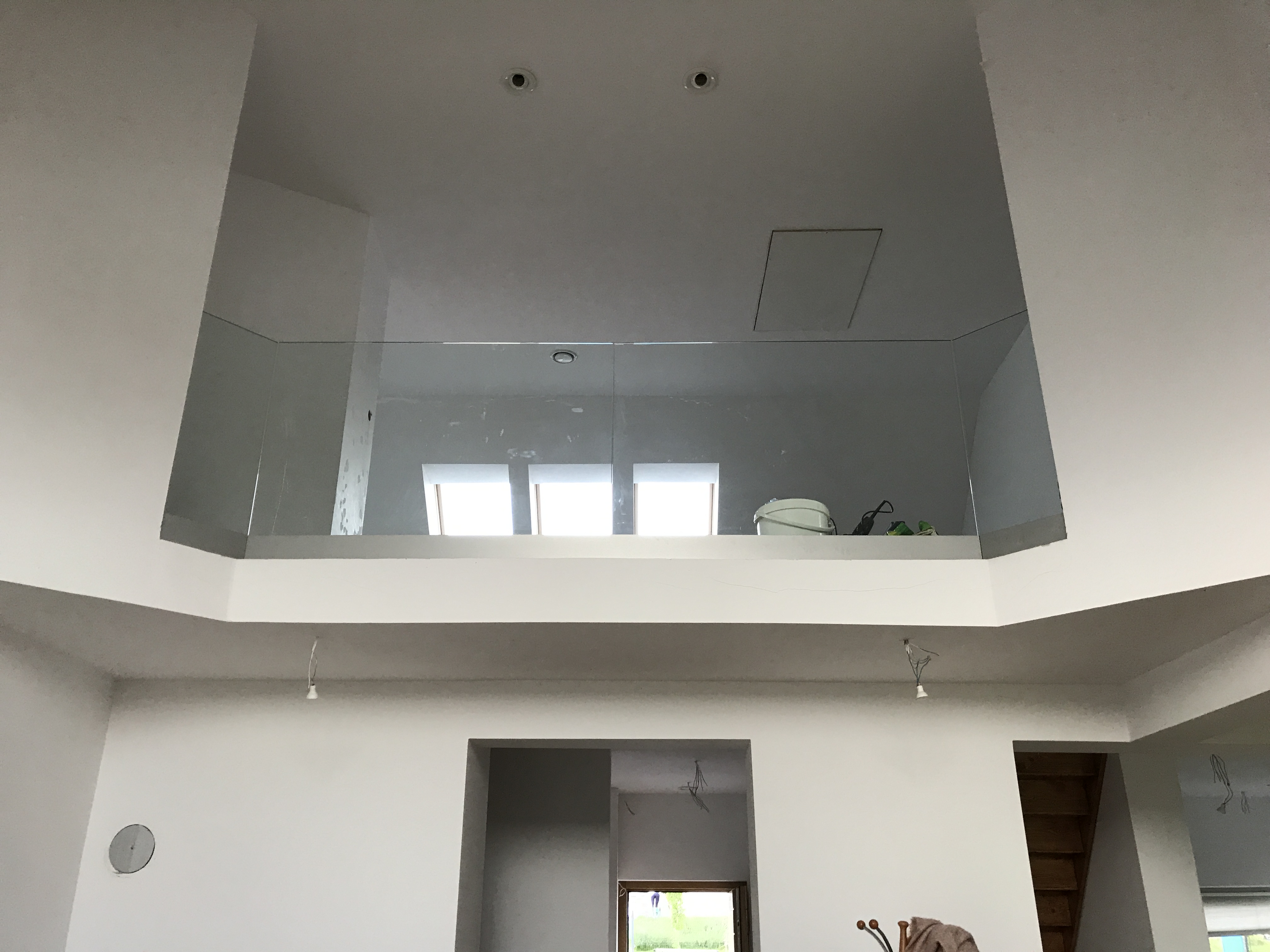 Szklana balustrada na antresoli w jasnym, minimalistycznym wnętrzu, widoczne okna dachowe i narzędzia malarskie, częściowo wykończony stan surowy.