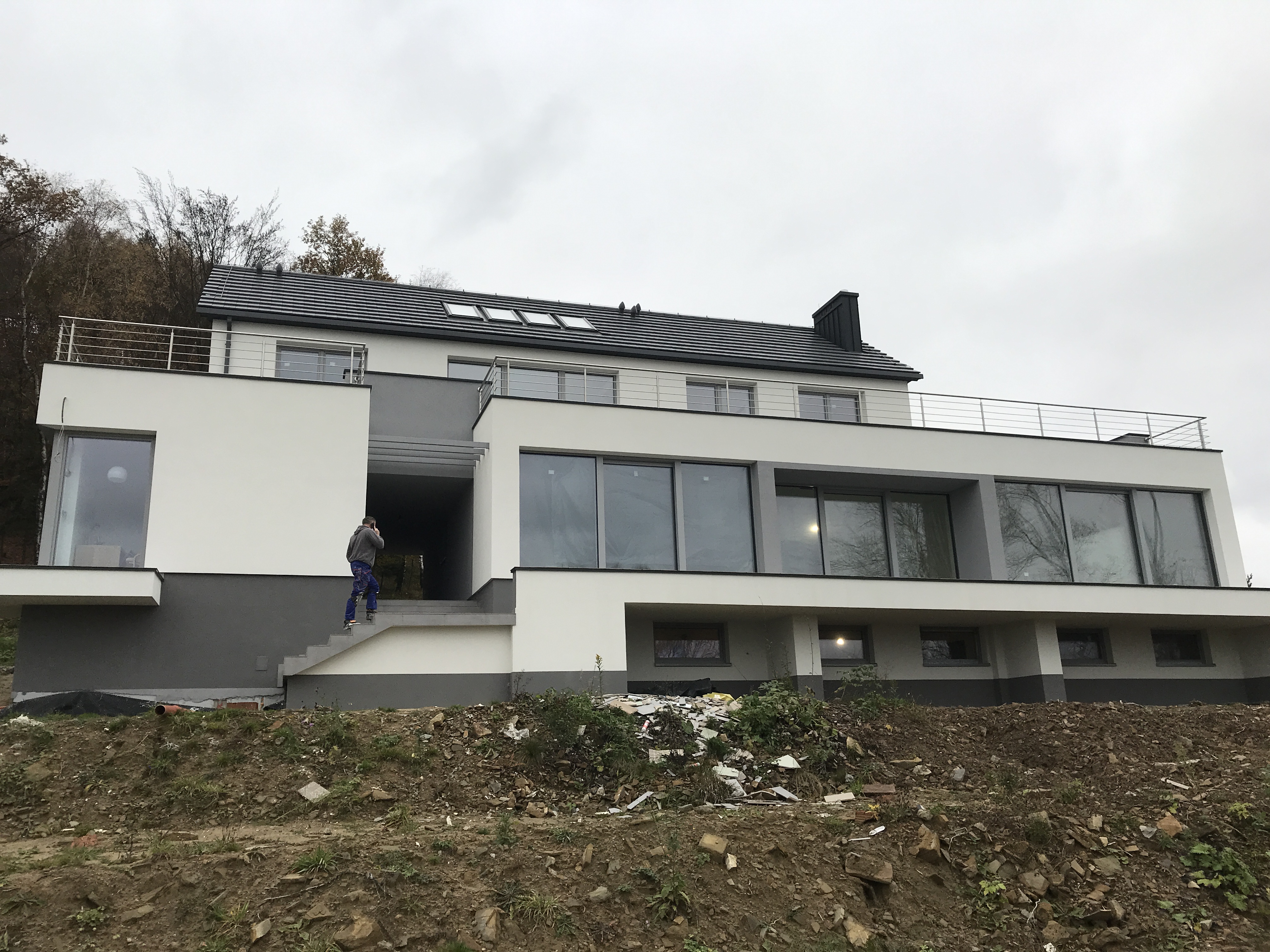 Nowoczesny dom z dużymi oknami, metalową balustradą na dachu i osobą wchodzącą po schodach zewnętrznych, teren wokół domu w trakcie prac wykończeniowych.