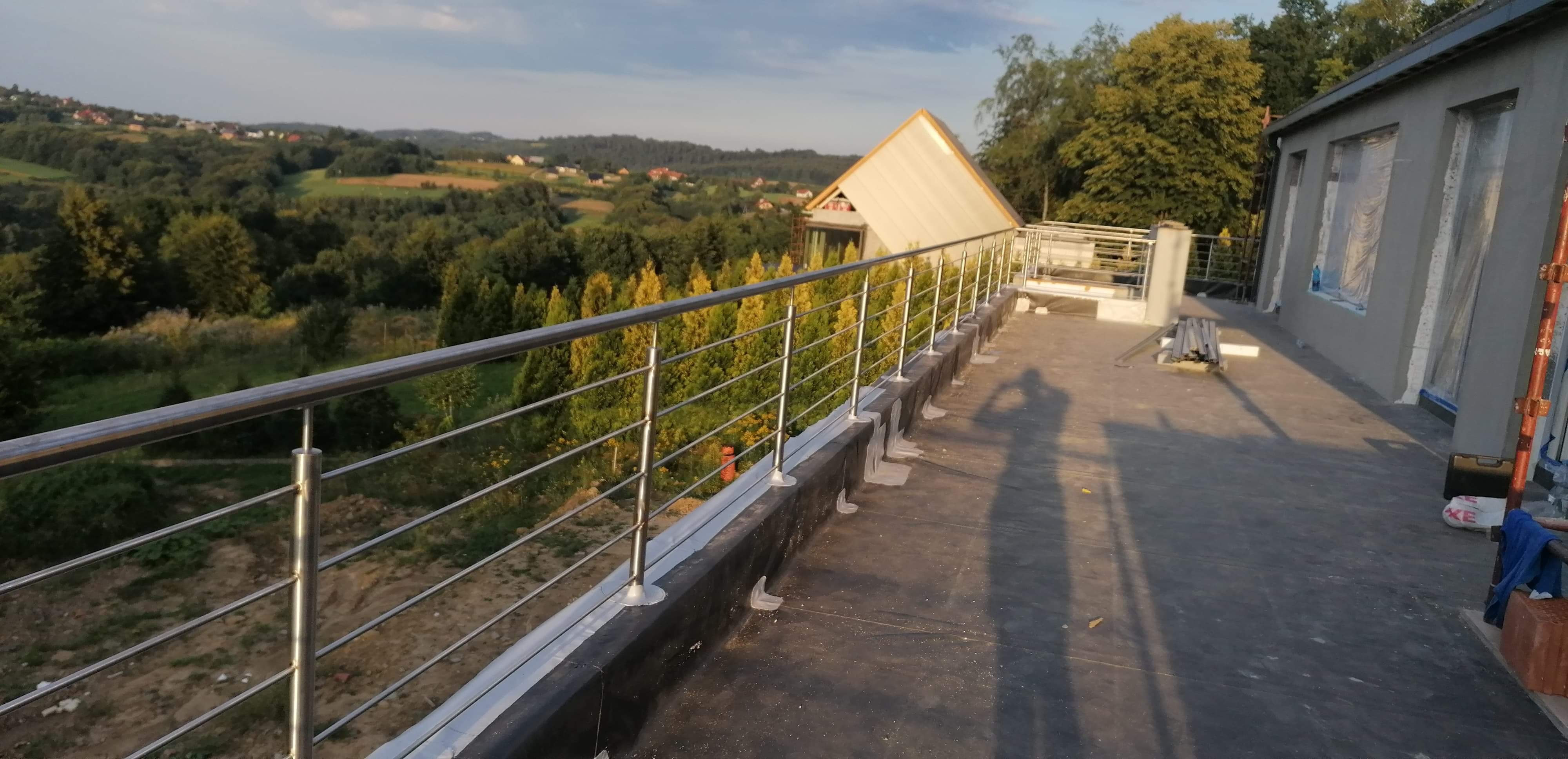 Metalowa balustrada na tarasie z widokiem na zielony krajobraz, cień osoby fotografującej widoczny na posadzce.