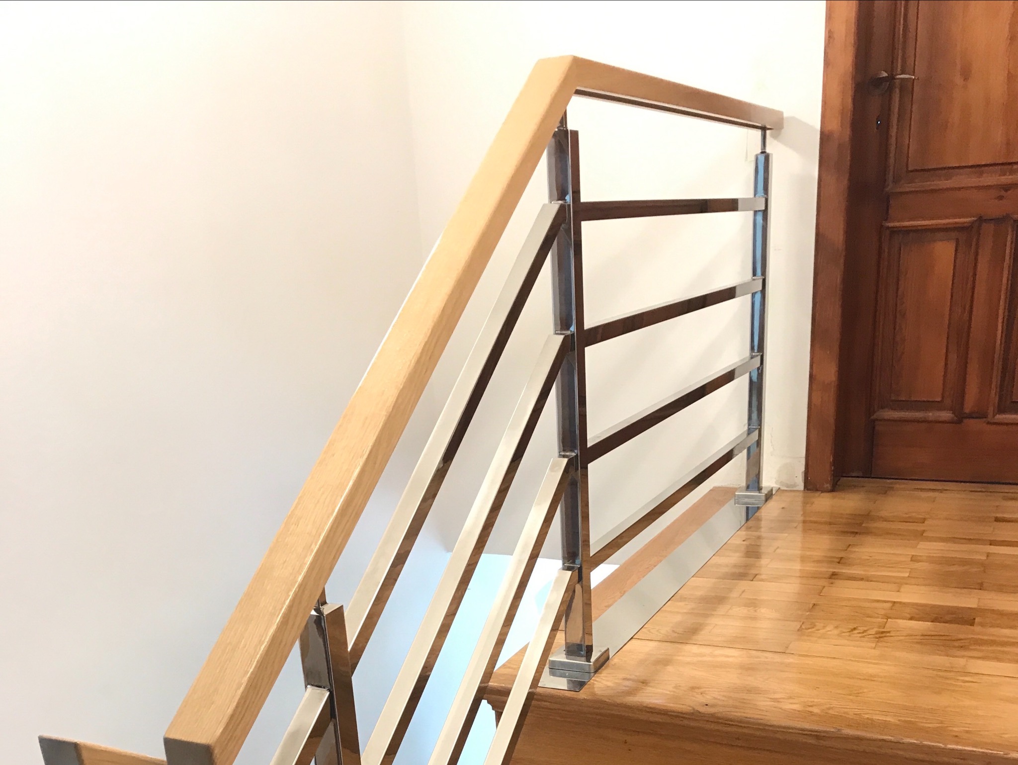 Nowoczesna balustrada schodowa z drewna i polerowanej stali nierdzewnej, widok z dołu schodów, parkiet dębowy na podłodze, brązowe drzwi w tle.