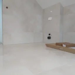 Jasne, błyszczące płytki podłogowe i ścienne w trakcie montażu w łazience, widoczne rury instalacyjne i kartony z materiałami.