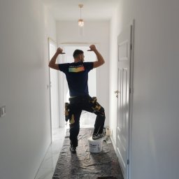 Dream house - Malarz w niebieskiej koszulce z napisem 'MAKE THE WORLD COLORFUL' świętuje sukces po pomalowaniu korytarza na biało, stojąc na wiadrze z farbą Flügger Dekso 5, z narzędziami przy pasie i podkładem...