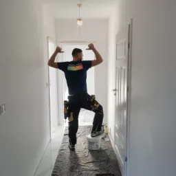 Malarz w niebieskiej koszulce z napisem 'MAKE THE WORLD COLORFUL' świętuje sukces po pomalowaniu korytarza na biało, stojąc na wiadrze z farbą Flügger Dekso 5, z narzędziami przy pasie i podkładem...