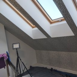 Dream house - Pokój na poddaszu po remoncie, ściany i skośny sufit wykończone tapetą w panterkę, widoczne okna dachowe, lampa na statywie i narzędzia pracy.