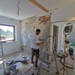 Dream house - Mężczyzna w trakcie tapetowania ściany w pokoju dziecięcym, widoczna mapa świata z motywami podróżniczymi, drabina, narzędzia i materiały budowlane, surowy stan wykończenia.