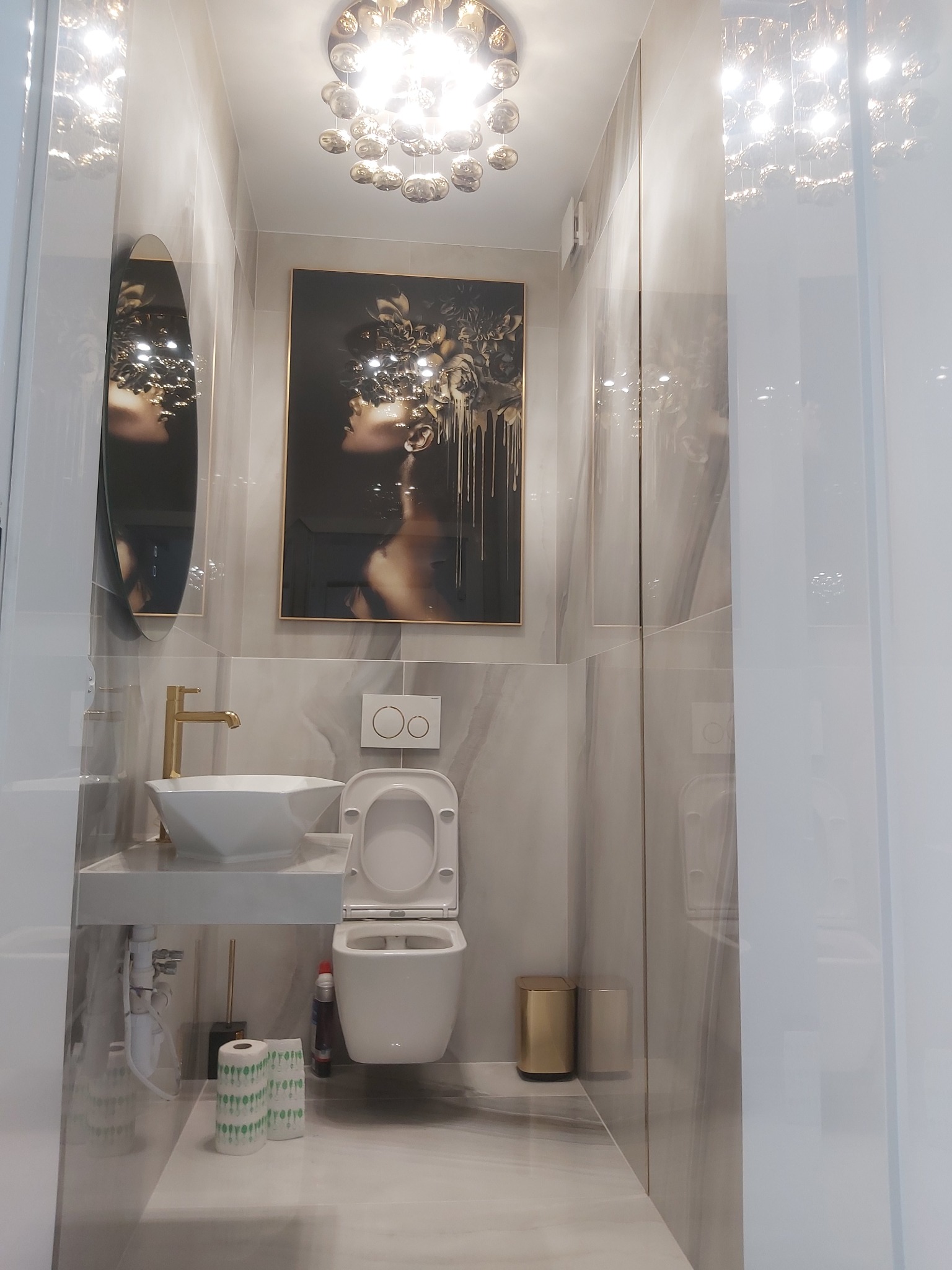 Nowoczesna łazienka w odcieniach szarości z marmurowymi płytkami, wiszącą toaletą, umywalką nablatową z złotą armaturą, okrągłym lustrem i dekoracyjnym obrazem kobiety z kwiatami.