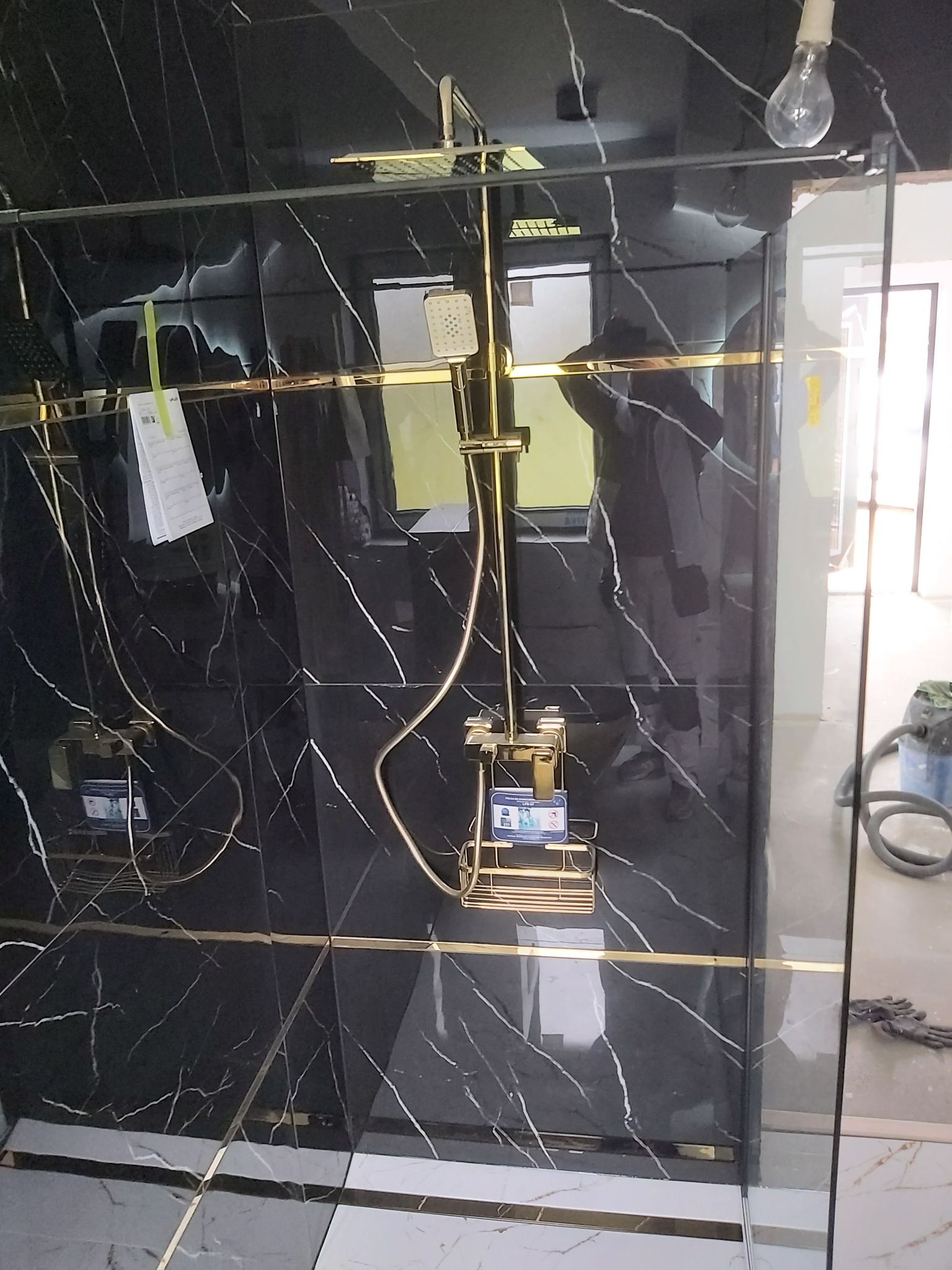 Kabina prysznicowa z czarnymi płytkami imitującymi marmur, złotymi akcentami i zamontowaną złotą armaturą prysznicową; widoczne narzędzia i odkurzacz w tle.