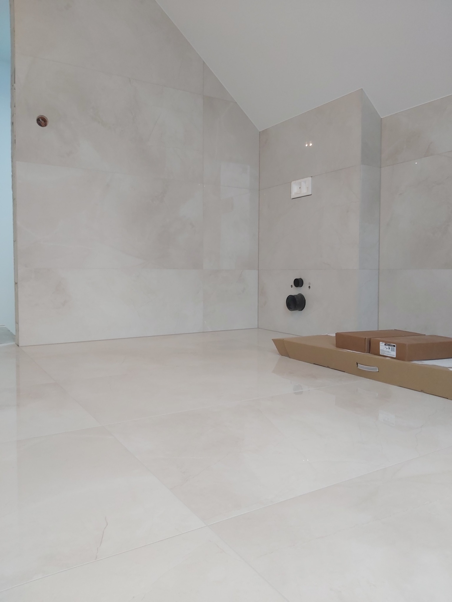 Jasne, błyszczące płytki podłogowe i ścienne w trakcie montażu w łazience, widoczne rury instalacyjne i kartony z materiałami.