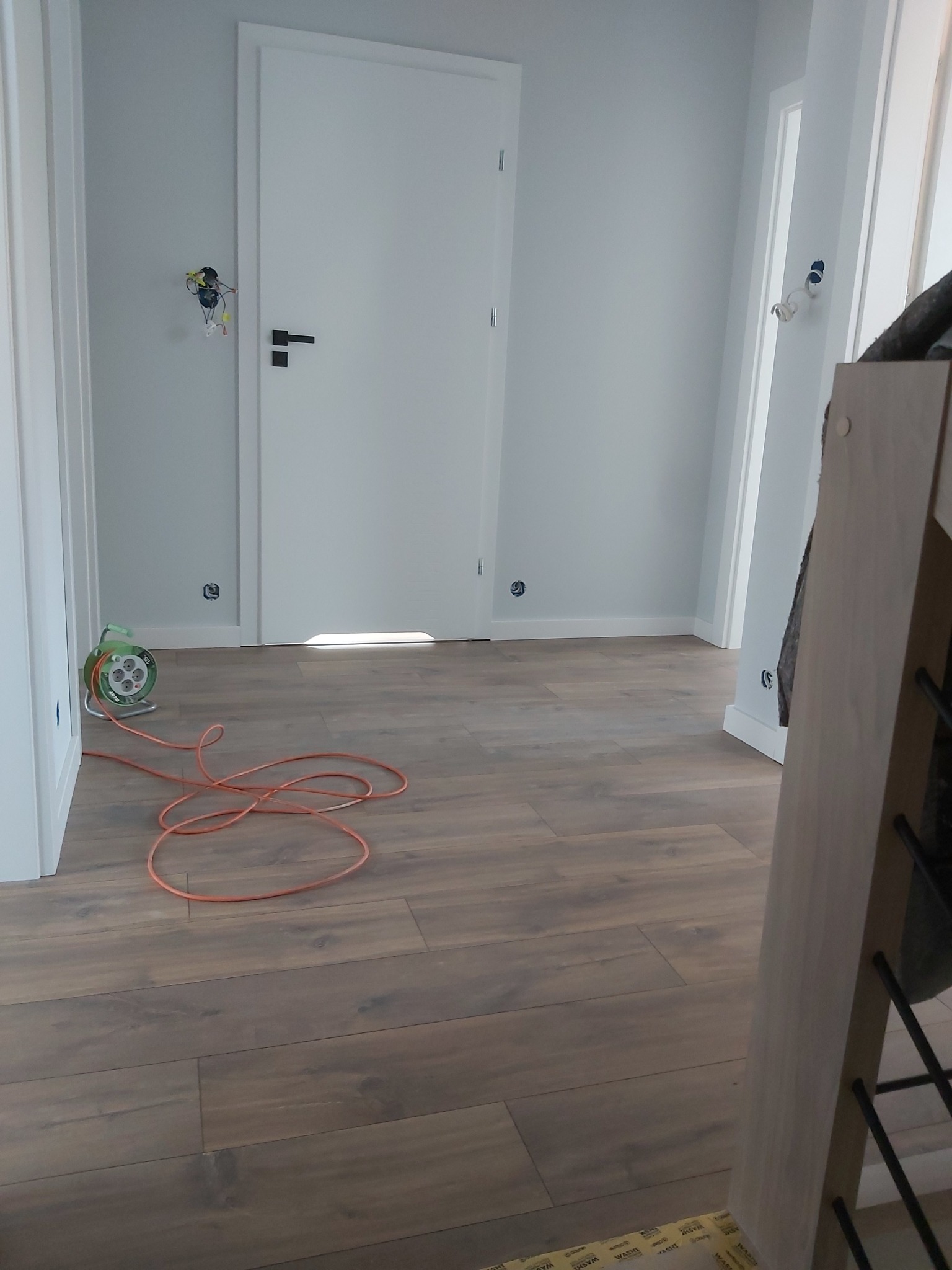 Nowa podłoga z paneli ułożona w jasnym pomieszczeniu, widoczny zwinięty przedłużacz i niedokończone gniazdko elektryczne bez osprzętu.