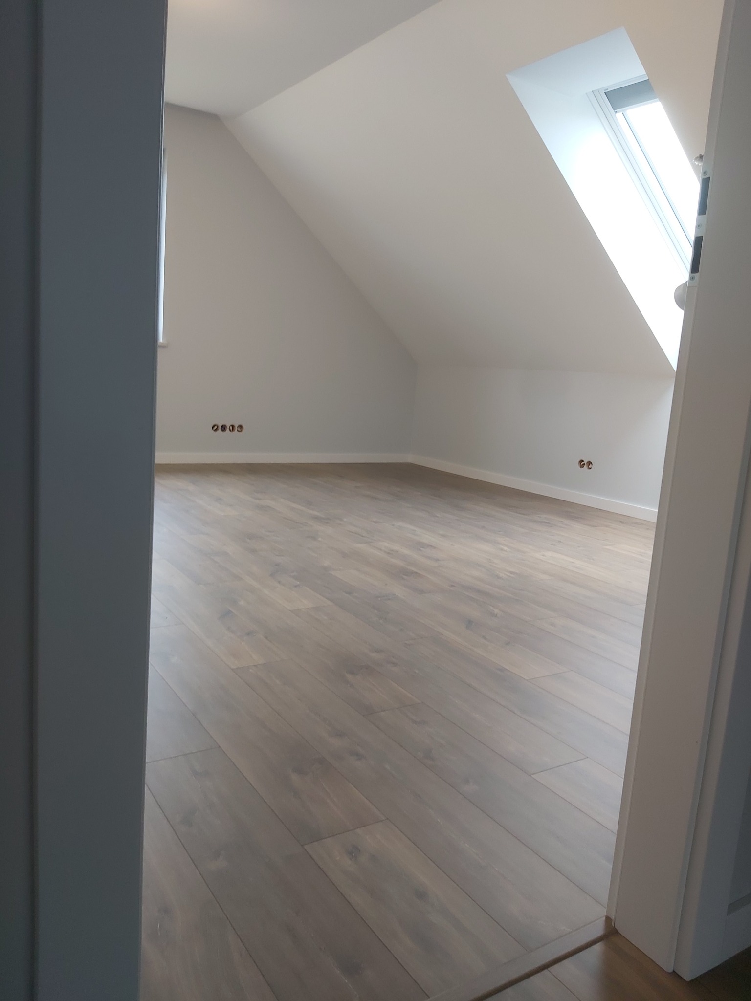 Poddasze z nową, jasną podłogą z paneli, widoczne gniazdka elektryczne i okno dachowe, ujęcie zza otwartych drzwi.