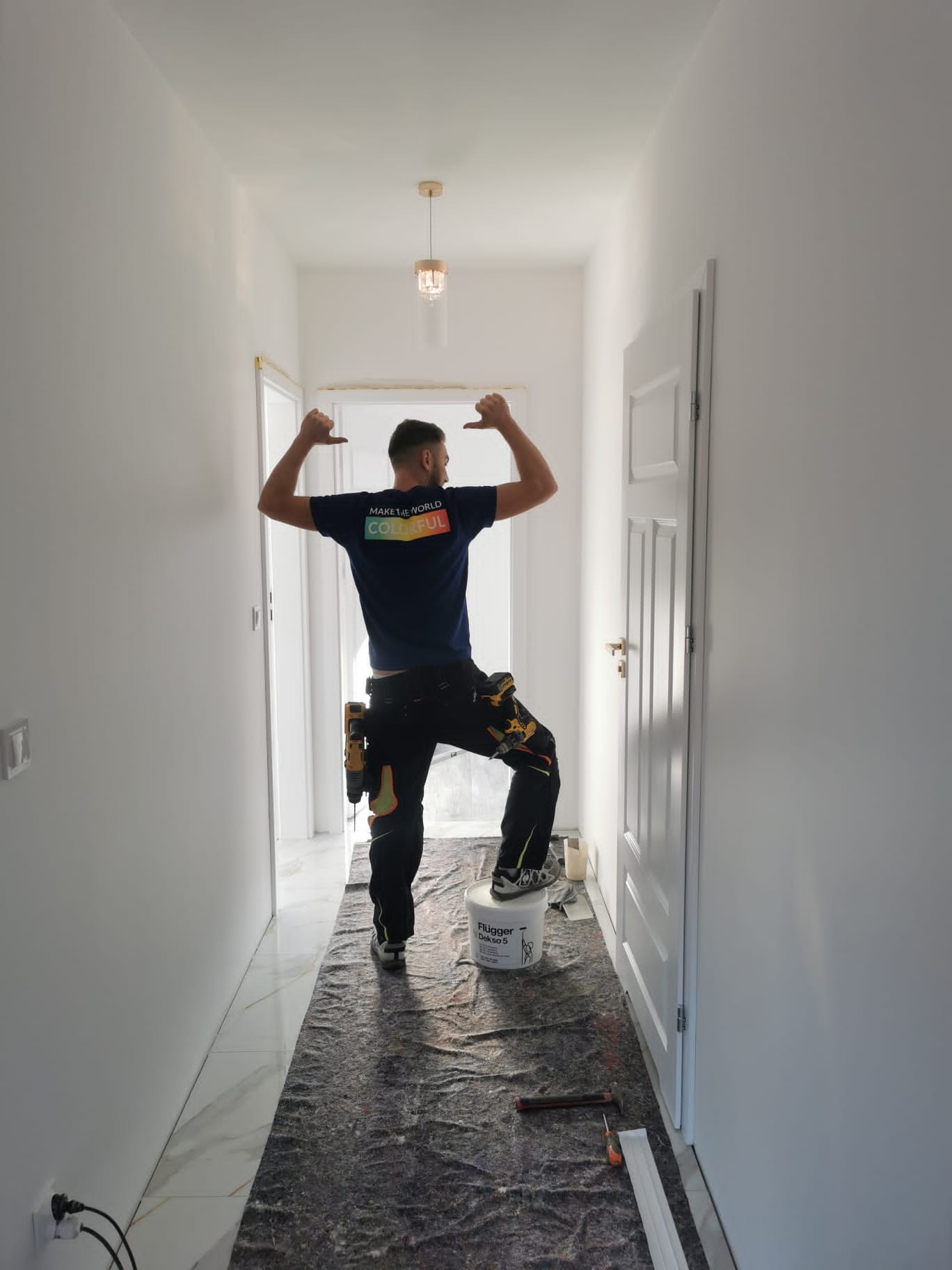 Malarz w niebieskiej koszulce z napisem 'MAKE THE WORLD COLORFUL' świętuje sukces po pomalowaniu korytarza na biało, stojąc na wiadrze z farbą Flügger Dekso 5, z narzędziami przy pasie i podkładem...