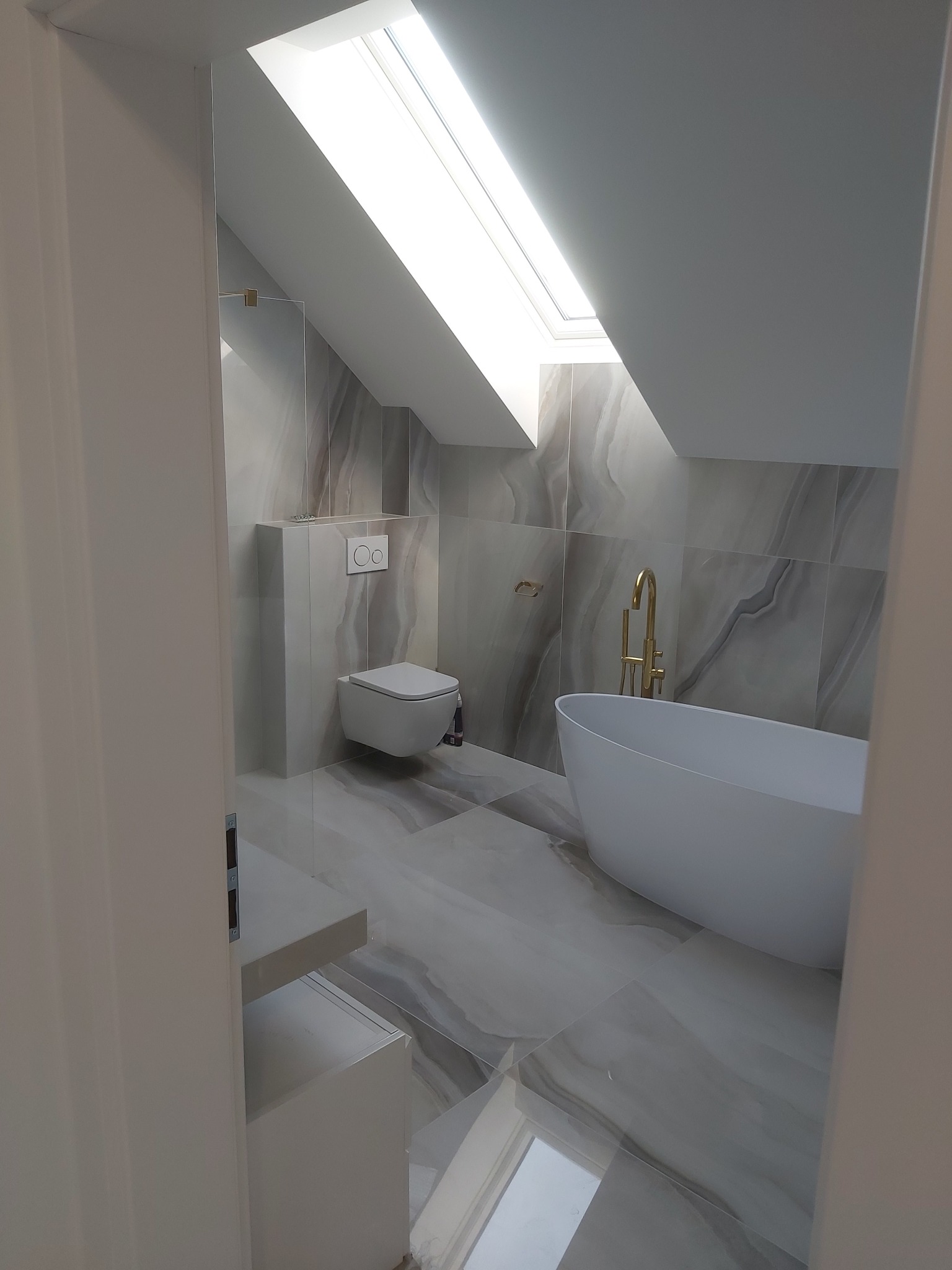 Nowoczesna łazienka na poddaszu z oknem dachowym, marmurowymi płytkami, wolnostojącą białą wanną i złotą armaturą.