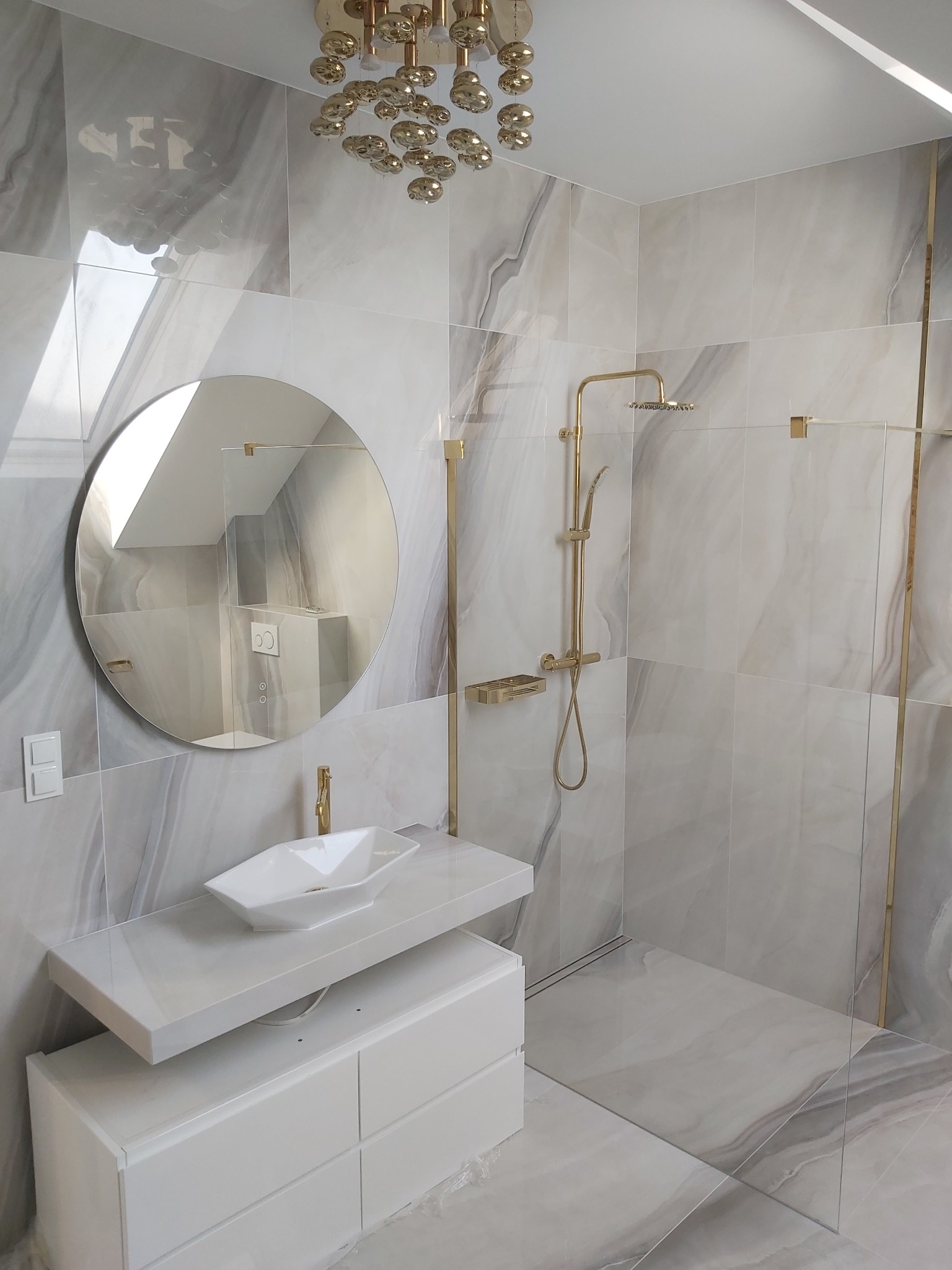 Elegancka łazienka wykończona marmurowymi płytkami z złotymi akcentami, okrągłe lustro na ścianie odbija skos dachu, biała szafka pod umywalką i nowoczesna złota armatura.