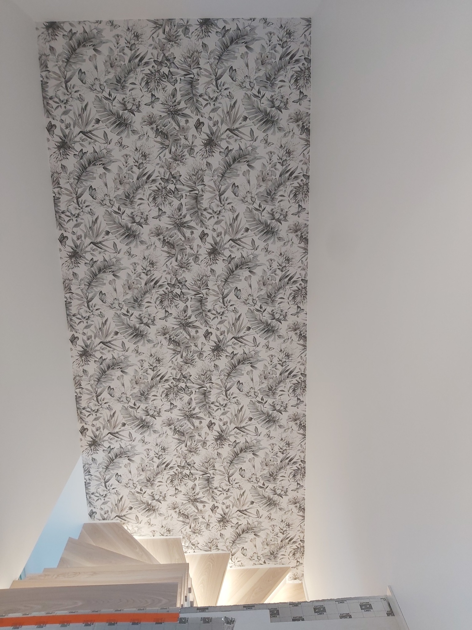 Ściana schodów z tapetą w motywy roślinne i motyle, widok z dołu, jasne schody drewniane w tle.