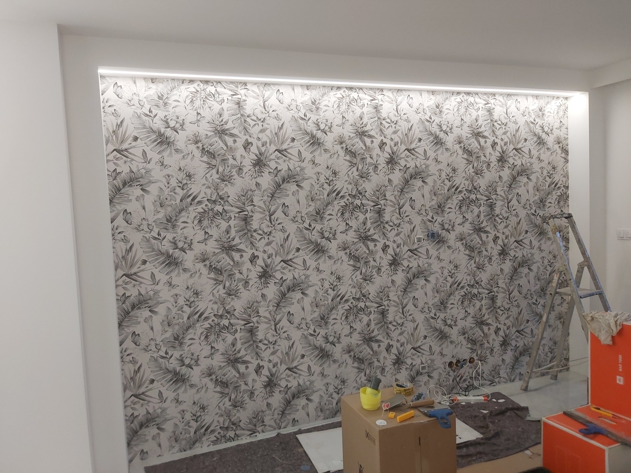 Ściana pokryta tapetą w motywy roślinne i motyle, oświetlona listwą LED, z widocznymi narzędziami i drabiną w trakcie prac wykończeniowych.