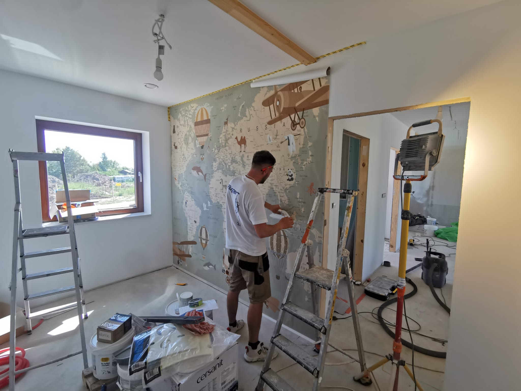 Mężczyzna w trakcie tapetowania ściany w pokoju dziecięcym, widoczna mapa świata z motywami podróżniczymi, drabina, narzędzia i materiały budowlane, surowy stan wykończenia.
