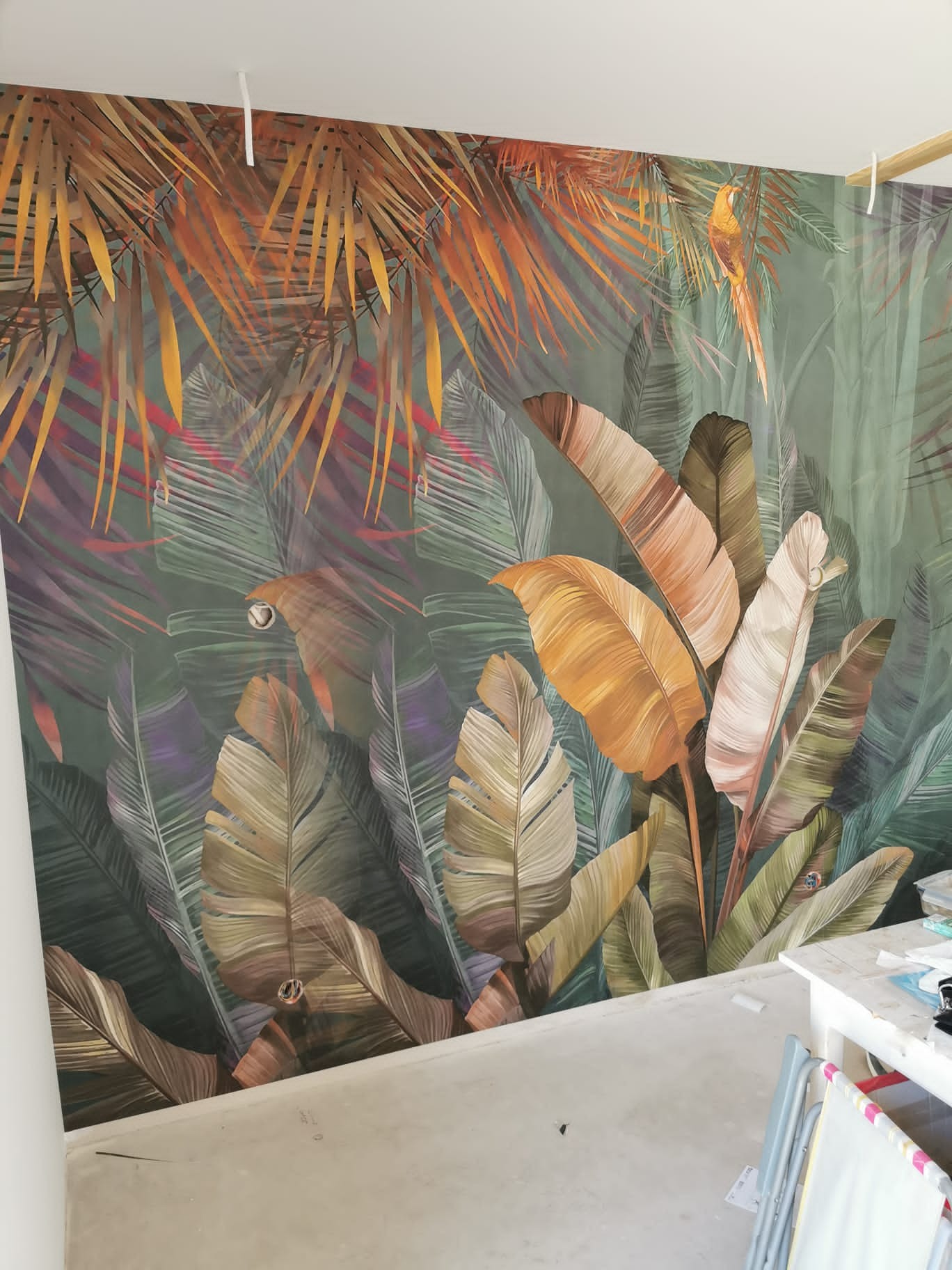 Ściana pokryta fototapetą z motywem tropikalnych liści bananowca i papug, widoczne elementy montażowe pod sufitem.